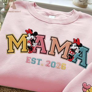 May include: Pink sweatshirt with the word "MAMA" in yellow, pink, and teal lettering, featuring Minnie Mouse graphics. The text "EST. 2026" is embroidered below. A matching pink garment is partially visible in the lower right corner.