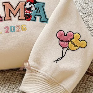 May include: Cream-colored sweatshirt with embroidered details. Features the word "MAMA" with a Minnie Mouse graphic, the year "2026", and two Mickey Mouse balloon designs with the names "ANNA" and "MATEO".