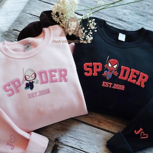 Może przedstawiać: Dwie bluzy, jedna różowa i jedna czarna, z haftowanymi wzorami "Spider". Różowa bluza ma grafikę Spider-Gwen, a czarna grafikę Spider-Mana. Obie zawierają "EST. 2018".