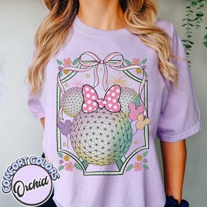 May include: A lilac Comfort Colors t-shirt with a graphic of a geometric sphere, a pink polka dot bow, Mickey Mouse ears, and floral details. The shirt has the words "Comfort Colors" and "Orchid" printed on it.