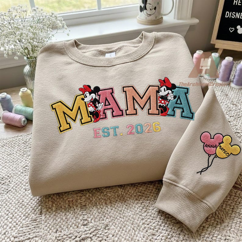 May include: Beige sweatshirt with the word "MAMA" in colorful embroidered letters, featuring Minnie Mouse graphics. The text "EST. 2026" is below the word "MAMA". A matching sleeve with Minnie Mouse balloon graphics is also visible.