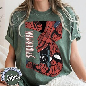 Puede incluir: Camiseta verde musgo con un dise&ntilde;o de Spider-Man rojo y negro. El dise&ntilde;o muestra las manos de Spider-Man sosteniendo una c&aacute;mara, con la palabra "SPIDER-MAN" impresa verticalmente. La camiseta tiene un logotipo de Comfort Colors.