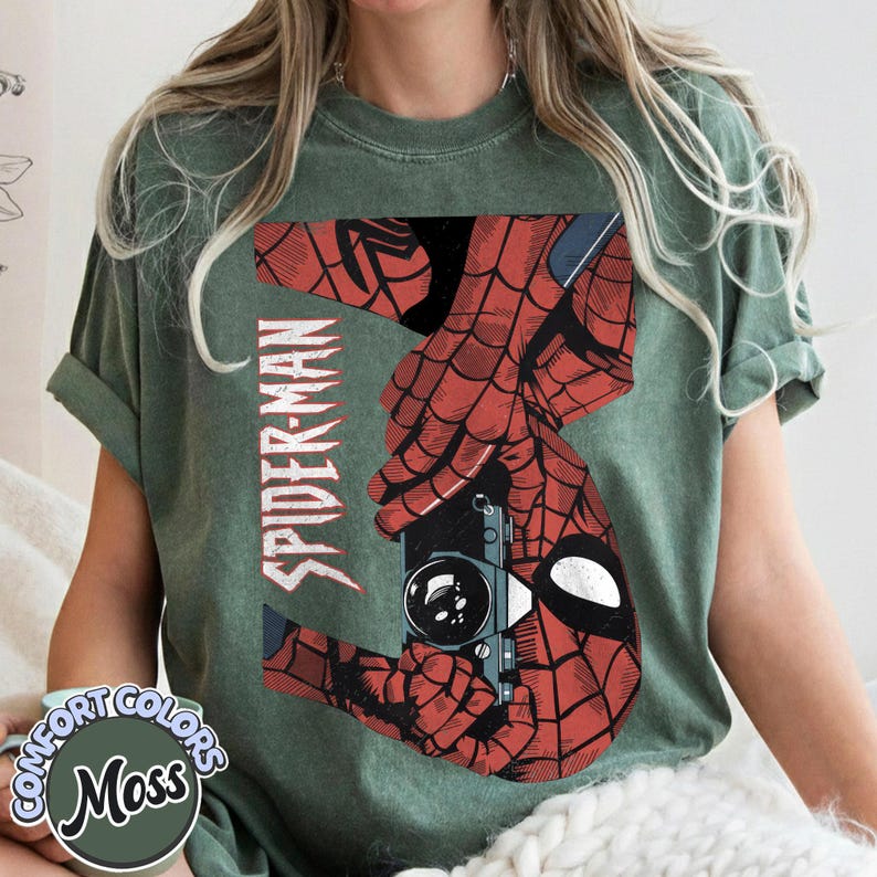 Puede incluir: Camiseta verde musgo con un dise&ntilde;o de Spider-Man rojo y negro. El dise&ntilde;o muestra las manos de Spider-Man sosteniendo una c&aacute;mara, con la palabra "SPIDER-MAN" impresa verticalmente. La camiseta tiene un logotipo de Comfort Colors.