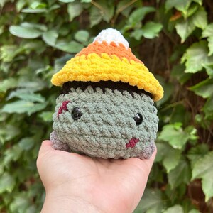 May include: A handmade crochet zombie doll with a candy corn hat. The doll is primarily green with black eyes, red stitches, and a small mouth. The hat is yellow, orange, and white. A unique Halloween decoration.