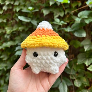 May include: A handmade, white, plush mushroom with a candy corn-colored cap. The cap features layers of white, orange, and yellow yarn. The mushroom has two black button eyes and a rounded body. The background is a green leafy plant.