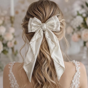 Könnte beinhalten: Eine große, cremeweiße Haarschleife aus Satin mit zwei langen, fließenden Bändern, die am Hinterkopf befestigt ist. Der Stoff hat eine glatte, glänzende Oberfläche und die Schleife ist in der Mitte mit einem passenden Knoten versehen, was für einen weichen Fall sorgt.
