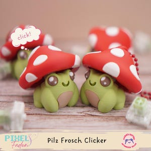 Könnte beinhalten: Zwei grüne Froschfiguren mit roten Pilzkappen und weißen Tupfen. Die Figuren haben große, ausdrucksstarke Augen und sind mit "Pilz Frosch Clicker" beschriftet. Eine Sprechblase sagt "click!".
