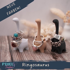 Könnte beinhalten: Drei Ringhalter in Dinosaurierform in Weiß, Beige und Schwarz, die jeweils mehrere Ringe tragen. Der weiße Dinosaurier hat einen weißen Hals, der beige einen beigen Hals und der schwarze einen schwarzen Hals. Der Text "NEUE FARBEN!" und "Ringosaurus" sind sichtbar.