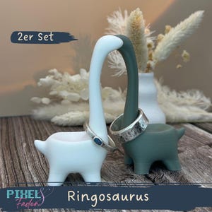 Könnte beinhalten: Zwei ringförmige Dinosaurierhalter, einer weiß und einer teal, die jeweils einen Silberring halten. Die Halter haben lange Hälse und kleine Körper. Der Text "2er Set" und "Ringosaurus" sind sichtbar.