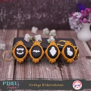Könnte beinhalten: Vier Halloween-Schlüsselanhänger mit goldfarbenen Rahmen und Schlüsselringen. Jeder Schlüsselanhänger hat ein schwarzes Oval mit einem weißen Design: Vampirzähne, eine Fledermaus, ein Geist und eine Katze. Der Text "Vintage Bilderrahmen" ist sichtbar.