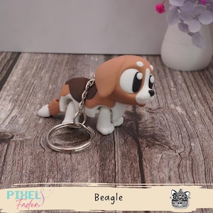 Könnte beinhalten: Ein Beagle-Schlüsselanhänger in Braun, Weiß und Beige. Der Hund hat große, ausdrucksstarke Augen und einen silbernen Schlüsselring. Das Wort "Beagle" ist darunter gedruckt.