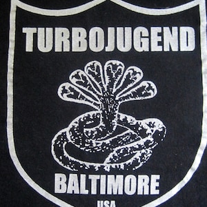 May include: A black t-shirt with a silver shield design. The shield features the text "TURBOJUGEND" and "BALTIMORE, USA" along with a graphic of a snake with multiple heads.