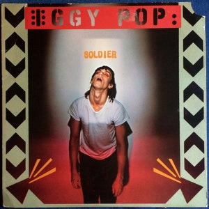 May include: A vintage record album cover featuring Iggy Pop. The cover is red and black with a white border. The album title "Soldier" is printed in yellow letters. Iggy Pop is wearing a white t-shirt and black pants. He is looking up with his mouth open.