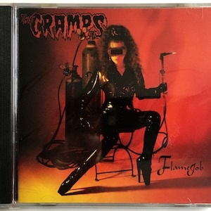 May include: A red CD case with a black spine. The front cover features a woman in a black leather outfit holding a welding torch. The text "The Cramps" and "Flame Job" are visible on the cover.