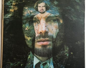 May include: Album cover art featuring a double exposure of a man's face superimposed over a forest scene. The man in the foreground has a beard and is wearing a patterned shirt and tie. The background features a second man and a forest.