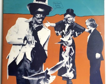 May include: Album cover art for Joni Mitchell's "Don Juan's Reckless Daughter." The artwork features three figures against an orange and teal background. One figure wears a white suit and hat, another a black dress with skulls, and a third a tuxedo.