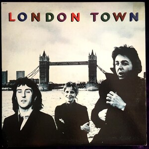 Pode incluir: Capa do álbum "London Town" com a Tower Bridge ao fundo. O título está em letras coloridas. Três pessoas estão em primeiro plano. O céu está nublado.