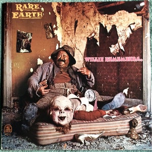 Puede incluir: Una portada de álbum de vinilo vintage con un hombre con sombrero y mono sentado en una cama con una muñeca acostada en su regazo. La portada del álbum se titula "Rare Earth" y "Willie Remembers..." en letras rosas.