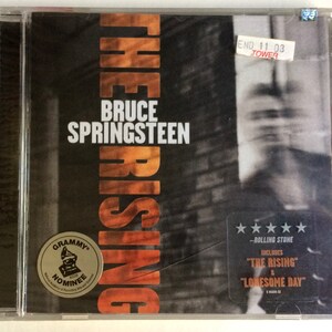 Puede incluir: Una caja de CD con una foto en blanco y negro de Bruce Springsteen en la parte delantera. El título del álbum es "The Rising" y el texto "Bruce Springsteen" está impreso en blanco. La caja de CD tiene una pegatina dorada que dice "Grammy Nominee 2003".