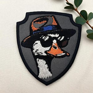 May include: Embroidered patch featuring a goose wearing sunglasses and a hat. The goose has a white body, black head, and an orange beak. The patch is shield-shaped with a gray background and a black border.