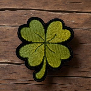 以下が含まれることがあります： 黒い縁取りのある緑色の四つ葉のクローバーパッチ。クローバーはさまざまな緑の色合いで刺繍されており、質感のある外観を作り出しています。パッチは木製の背景に配置され、詳細なステッチを強調しています。