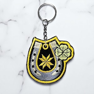 Peut inclure: Porte-clés brodé avec un fer à cheval et un trèfle à quatre feuilles. Le fer à cheval est noir et argenté, avec un bord jaune. Le trèfle est vert clair et le porte-clés a un anneau argenté.