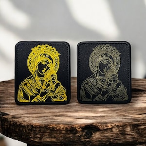 May include: Two square black patches with a gold religious icon design. The icon shows a woman holding a child, encircled by a halo. The patches are on a wooden surface, highlighting the detailed embroidery.