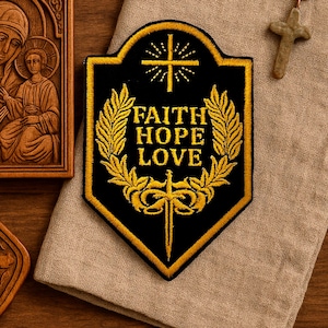 Puede incluir: Un parche bordado en negro y dorado con las palabras "FAITH HOPE LOVE". El parche presenta una cruz, espigas de trigo y un diseño de espada. El parche está sobre una tela beige.