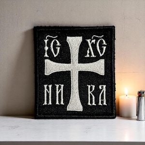 Könnte beinhalten: Schwarzer, bestickter Aufnäher mit einem weißen Kreuz und stilisiertem Text. Der quadratische Aufnäher ist wahrscheinlich für Kleidung oder Accessoires mit religiösem Thema. Eine brennende weiße Kerze und ein silbernes Feuerzeug liegen daneben.