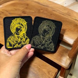 Op de afbeelding: Twee vierkante zwarte patches met goud geborduurde religieuze iconen van een vrouw en een kind. De iconen zijn omgeven door gouden halo's. De patches worden gepresenteerd tegen een houten achtergrond.
