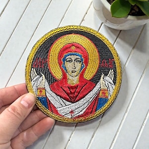 Op de afbeelding: Cirkelvormige geborduurde patch met een religieus icoon in een rode jurk en blauwe hoofdbedekking, omringd door een gouden halo. De patch heeft een zwarte achtergrond, gouden rand en rode tekst.