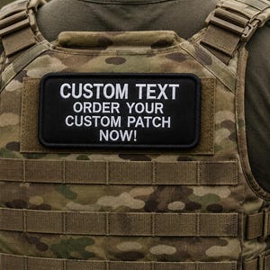 May include: A rectangular black patch with white embroidered text reading CUSTOM TEXT ORDER YOUR CUSTOM PATCH NOW! attached to a camouflage tactical vest. The patch features a clean border and is centered on the upper back section of the vest.