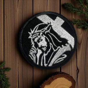 Puede incluir: Parche bordado redondo negro con una representación plateada de Jesús con una corona de espinas y una gran cruz en el fondo. El parche está sobre un fondo de madera con una ramita verde.