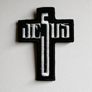 Op de afbeelding: Zwart geborduurde patch in de vorm van een kruis. Het woord "JESUS" is in witte blokletters op de bovenkant van het kruis geborduurd. Een verticale witte lijn loopt door het midden van het kruis.