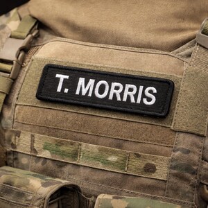 Op de afbeelding: Een zwart rechthoekig embleem met de tekst "T. MORRIS" in het wit, bevestigd op een beige en camouflage tactisch vest. Het vest heeft meerdere riemen en zakken, typerend voor militaire of tactische uitrusting.