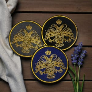 Op de afbeelding: Drie ronde geborduurde patches met een gouden dubbelkoppige adelaar ontwerp en een kroon. De patches hebben gouden randen en zijn geplaatst tegen zwarte, donkergrijze en blauwe achtergronden. Een takje lavendel staat op de achtergrond.