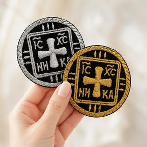 Puede incluir: Dos parches bordados circulares con fondo negro, una cruz central y las letras griegas IC XC NI KA. Un parche presenta detalles en hilo metálico plateado, mientras que el otro está bordado con hilo metálico dorado.