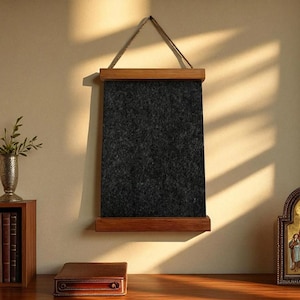 Puede incluir: Un tablero de fieltro gris oscuro con rieles de madera en la parte superior e inferior, suspendido por un cordón de fibra natural. El tablero está colgado en una pared de color claro, con la luz del sol que lo atraviesa. El tema general es la decoración del hogar.