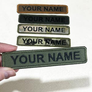 May include: Rectangular embroidered patches in various colors, including brown, black, and green, each displaying the text "YOUR NAME" in a contrasting color. The patches have a military-style aesthetic, with some featuring camouflage patterns.