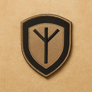 May include: Embroidered shield-shaped patch with a black border and a tan centre. The centre features a black rune symbol. The patch is designed to be sewn or ironed onto clothing or accessories.