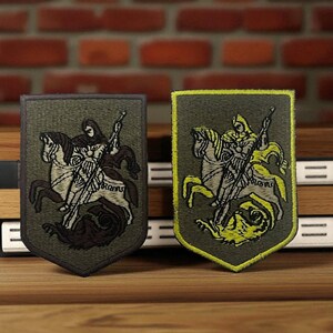 Può includere: Due toppe ricamate raffiguranti un cavaliere a cavallo che uccide un drago. Le toppe hanno la forma di uno scudo con uno sfondo verde scuro e disegni dettagliati in marrone e beige. Una toppa ha un bordo giallo.