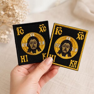 以下が含まれることがあります： 黒い正方形の刺繍ワッペン2枚。中央には暗い髪と髭の顔が描かれた円形の金色のアイコンがあり、白と金の十字のディテールと、四隅に金色の糸で刺繍されたギリシャ文字のIC XC NI KAがデザインされています。
