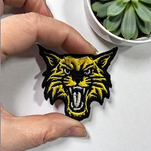 May include: Embroidered patch featuring a snarling yellow and black wildcat. The patch has a detailed design with visible teeth and sharp features. The patch is held between fingers against a white background.