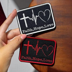 Puede incluir: Dos parches rectangulares bordados. Uno es negro con una cruz blanca, una línea de latidos y un corazón, con las palabras "Faith, Hope, Love". El otro es negro con una cruz roja, una línea de latidos y un corazón, con las mismas palabras.
