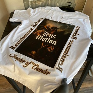 May include: White t-shirt featuring a Renaissance-style graphic with the words "Zeus Motion" in the center. The shirt also has the phrases "Renaissance Reborn" and "Chapter of Thunder" printed around the graphic in a brown color.