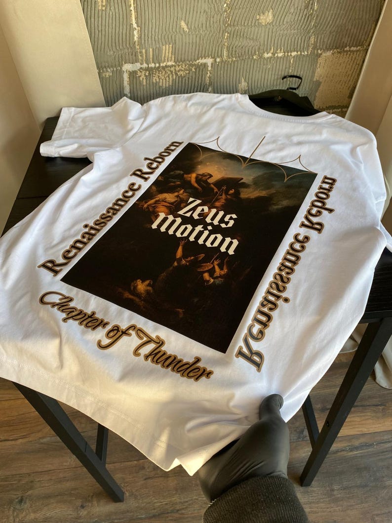 May include: White t-shirt featuring a Renaissance-style graphic with the words "Zeus Motion" in the center. The shirt also has the phrases "Renaissance Reborn" and "Chapter of Thunder" printed around the graphic in a brown color.