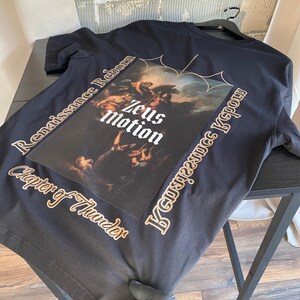 May include: Black t-shirt featuring a Renaissance-style graphic with the text "Zeus Motion." The shirt also includes the phrases "Renaissance Reborn" and "Chapter of Thunder" in a gold color. The shirt is displayed on a black table.