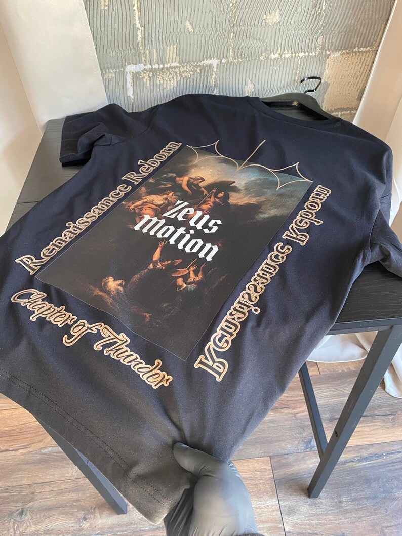 May include: Black t-shirt featuring a Renaissance-style graphic with the text "Zeus Motion." The shirt also includes the phrases "Renaissance Reborn" and "Chapter of Thunder" in a gold color. The shirt is displayed on a black table.
