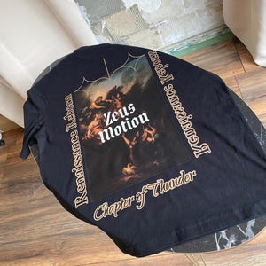 May include: Black long-sleeve shirt featuring a Renaissance-style painting with the text "Zeus Motion" in white. The shirt also has the text "Renaissance Reborn" and "Chapter of Thunder" in a gold color.