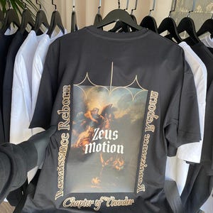 May include: Black t-shirt with a Renaissance-style graphic featuring the text "Zeus Motion" and "Chapter of Thunder." The design includes an image of a classical painting with figures. The shirt is hanging on a rack with other black and white shirts.